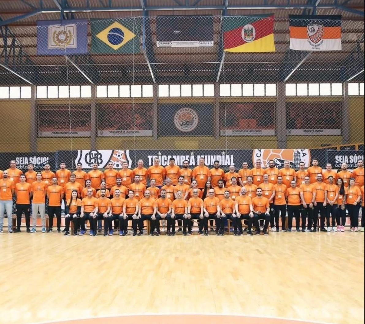 Turma do Curso de Treinadores, todos com camiseta laranja, posando no ginásio da ACBF em Carlos Barbosa, sob as bandeiras do Brasil e do Rio Grande do Sul